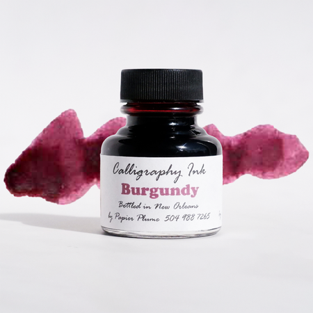 Bottled Papier Plume Burgundy calligraphy ink in a glass jar with black calligraphy pens on a white background.