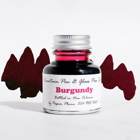 Papier Plume burgundy fountain pen ink bottle with ink flowing into a glass jar, showcasing rich burgundy color and elegant design.