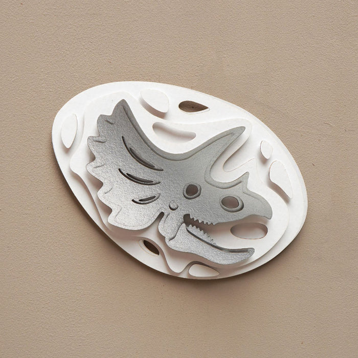 Silver paper fossil art of a Triceratops by Assembli, showcasing detailed prehistoric dinosaur design on textured paper.