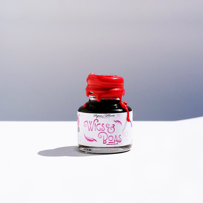 Papier Plume New Orleans Collection fountain pen ink bottle with vibrant ink inside, displayed alongside colorful feather boas.